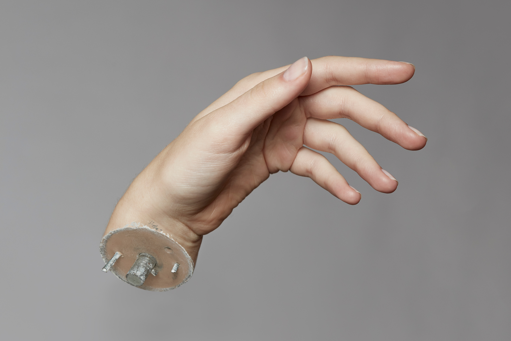 A mannequin hand against a neutral background, cut off at the wrist and attached to a flat circular metal plate with bolts, giving it a mechanical, prosthetic-like appearance.